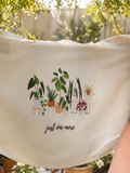Beige tote bag with plant illustration and text 'just one more' in a garden setting