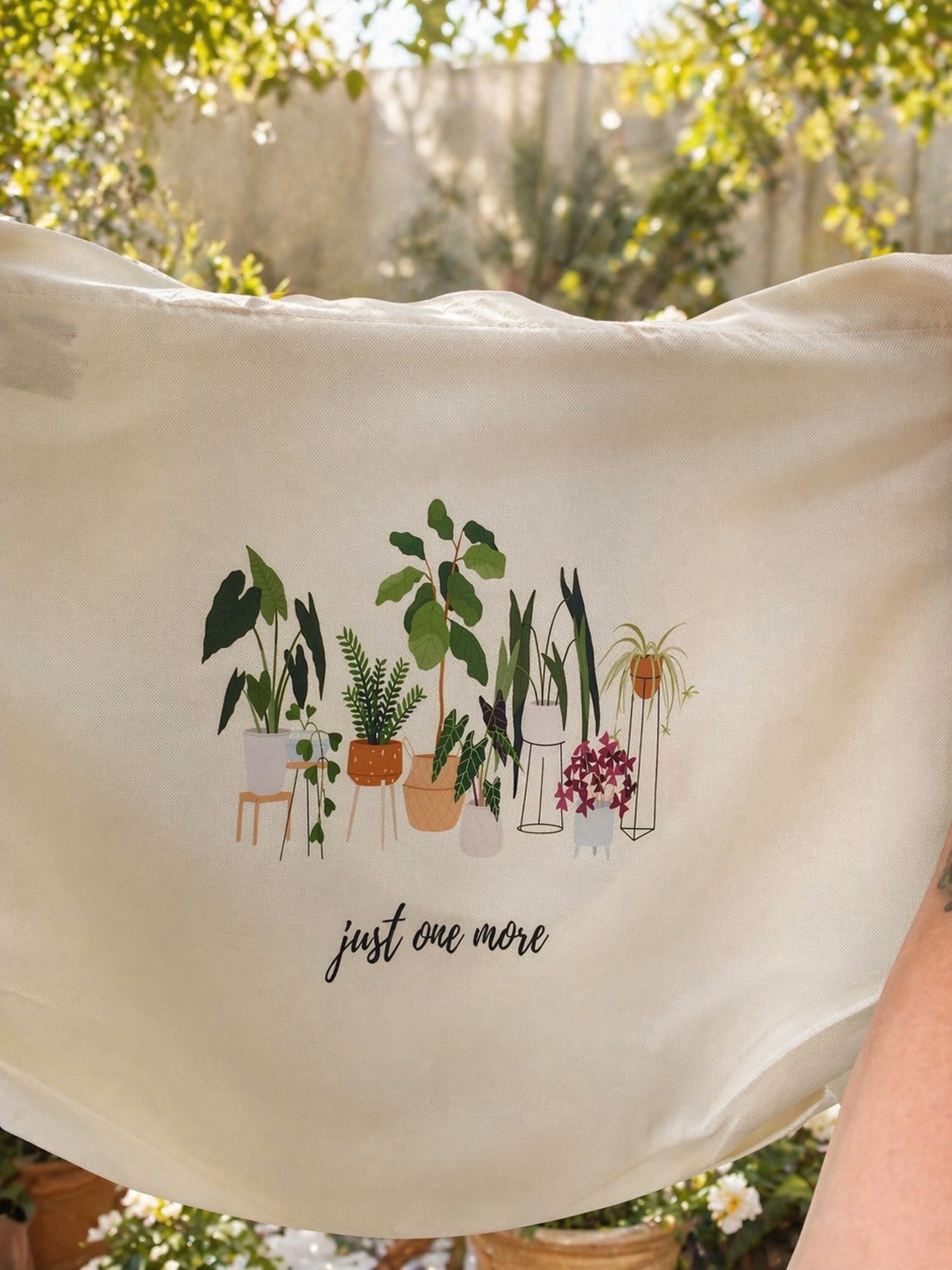 Beige tote bag with plant illustration and text 'just one more' in a garden setting